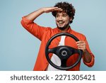 Small photo of Young fun Indian man wear orange red shirt white t-shirt hold steering wheel driving car look far away distance isolated on plain pastel light blue cyan background studio portrait. Lifestyle concept