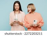 Small photo of Curious nosy elder parent mom with young adult daughter two women together wearing casual clothes holding in hand use mobile cell phone peep isolated on plain blue cyan background. Family day concept