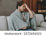 Side view young Indian man wapped in plaid hold head put hand forehead sufer from hangover headache sits on grey sofa couch stay at home hotel flat spend free spare time in living room. Lounge concept
