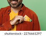 Close up smiling cheerful fun toothy bearded happy man he wears casual clothes t-shirt red shirt hold in hand put mock up of credit bank card in pocket isolated on plain pastel light green background