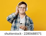 Young satisfied man with down syndrome wear glasses casual clothes headphones listen to music use mobile cell phone isolated on pastel plain yellow color background. Genetic disease world day concept