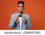 Young fun rich employee business man corporate lawyer wear classic formal grey suit shirt glasses work in office hold in hand mock up of credit bank card isolated on plain red orange background studio