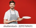 Small photo of Young waiter smiling fun male housewife housekeeper chef cook baker man wear grey apron hold point hand on empty plate with workspace area isolated on plain red background studio. Cooking food concept