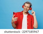 Small photo of Young cheerful happy fun vivid man of African American ethnicity 20s he wearing red shirt headphones listen music sing song use mobile cell phone isolated on plain pastel light blue cyan background