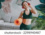 Young calm strong sporty athletic fitness trainer instructor woman wear green tracksuit headphones listen music hold in hand yoga mat training do exercises at home gym indoor. Workout sport concept