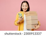 Small photo of Young surprised happy woman of Asian ethnicity wears yellow shirt white t-shirt hold stack cardboard blank boxes use mobile cell phone isolated on plain pastel light pink background studio portrait