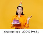 Happy fun surprised shocked young woman wears casual clothes hat celebrating hold in hand purple cake with candles spread arm isolated on plain yellow background. Birthday 8 14 holiday party concept