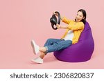 Full body fun young woman of Asian ethnicity wear yellow shirt white t-shirt sit in bag chair hold steering wheel driving car isolated on plain pastel light pink background studio. Lifestyle concept