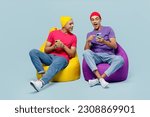 Full body cheerful happy happy young couple two friend men wear casual clothes together sit in bag chair hold in hand use mobile cell phone isolated on pastel plain light blue cyan background studio