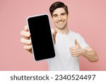 Small photo of Young man wear white t-shirt casual clothes hold in hand using close up mobile cell phone in blue case with blank screen workspace area show thumb up isolated on plain pastel pink background studio