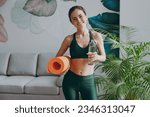 Young fun strong sporty athletic fitness trainer instructor woman wearing green tracksuit hold in hand yoga mat drink water training do exercises at home gym indoor. Workout sport motivation concept