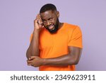 Small photo of Preoccupied worried young man of African American ethnicity wearing basic empty blank orange t-shirt stand put hand on head looking camera isolated on pastel violet colour background studio portrait