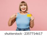 Small photo of Elderly smiling cheerful blonde woman 50s years old she wear blue undershirt casual clothes hold in hand point on mock up of credit bank card isolated on plain light pink background. Lifestyle concept