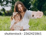 Smiling happy young woman in white clothes do selfie shot on mobile cell phone v-sign gesture child baby girl 5-6 years old Mommy rest with little kid daughter outdoor together. Love family concept.