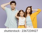 Small photo of Young shocked sad amazed parents mom dad with child kid daughter girl 6 years old wear blue yellow casual clothes look camera hold scratch head isolated on plain purple background. Family day concept