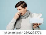 Small photo of Young ill sick man wear gray sweater scarf sneezing hold box of paper napkins sneeze isolated on plain blue background studio. Healthy lifestyle disease virus treatment cold season recovery concept