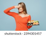 Small photo of Young dissatisfied sad woman wear orange casual clothes close nose sniff bad smell hold eat raw fresh sushi roll served on black plate Japanese food isolated on plain blue background studio portrait