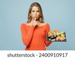 Small photo of Young secret woman wear orange casual clothes say hush be quiet, finger lips shhh gesture hold eat raw fresh makizushi sushi roll served on black plate Japanese food isolated on plain blue background