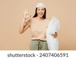 Small photo of Young calm Latin woman she wears pyjamas jam sleep eye mask rest relax at home hold pillow show ok okay gesture wink isolated on plain pastel light beige background studio. Good mood night nap concept