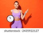 Young fitness trainer instructor sporty woman sportsman wear purple top clothes spend time in home gym hold clock point finger aside isolated on plain orange background. Workout sport fit abs concept