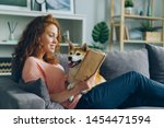 Pretty student young woman is reading book in cozy apartment smiling and petting adorable dog sitting on comfy couch at home. Animals and hobby concept.