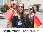 Stylish slim ladies wearing trench-coat and sunglasses walking with colorful shopping bags near the store during shopping process, concept of consumerism, sale, rich life