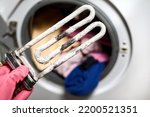 A man holds in his hand a burnt-out heating element of a washing machine against the background of a washing machine
