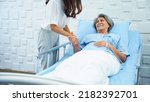 Small photo of Young woman lifting a railing beside the bed and holding his sick mother's hand to get saline on the bed in the hospital room. Daughter encouraging mother lying sick in the patient room.