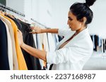 Small photo of Pretty young woman standing in front of a hanger and trying to choose clothes for work or outing. Selection of wardrobe, stylist, shopping.