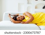 Beautiful curvy oversize young smiling  african american woman in a yellow dress lies on a sofa at home. Body positive stylish smiling female have relax time. Body acceptance and diversity concept