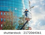 Small photo of Man in cradle painting lamppost. Worker in aerial platform, man paint street light pole at height, renovation works. Worker in lift bucket at height, risky job. Paint and renovate street light