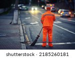 Small photo of Man with broom clean city road and roadside at night. Worker cleans roads, streets, footpaths with broom, night work. Janitor cleanup city from garbage. Municipal worker in uniform sweeping street