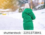Woman walking alone on snowy city street during blizzard. Woman in green down jacket and knitted white hat, warm clothing in cold weather. Fashionable female walk through snowfall. 