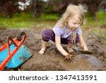 Small photo of Funny little girl playing in a large wet mud puddle on sunny summer day. Child getting dirty while digging in muddy soil. Messy games outdoors.