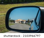 Side-view mirror on driver's side of car reflects perspective of suburban street with single-family houses under construction along one side on a sunny morning in southwest Florida