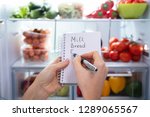 Hand Making List Of Food On Spiral Notepad In Front Of An Open Refrigerator