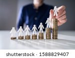 Human Hand Placing Small Human Figure On Increasing Stacked Coins Over Wooden Desk