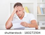Businessman sitting in an office reacting in shock to the contents of a letter that he is reading raising his hand to his mouth