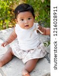 Small photo of Pretty little girl sitting unsupported on a stone seat surrounded by green plants, of mixed ethnic race and barefoot.