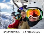Four positive young friend snowboarders in motion by cable car climb to top of mountain on winter resort of Rosa Khutor. Reflection of photographer in ski mask