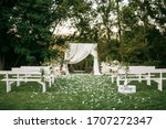 Wedding ceremony aisle with an arch made of chiffon white textile and flowers and long white wooden benches on green grass. Backyard wedding venue.