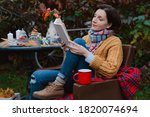 Small photo of young woman brown coat sits chair at table reading book with plaid thrown over her head open air against autumn reddened foliage. autumn fashion season. Rest, tranquility, backyard, patio, melancholy