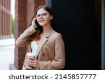 Small photo of Close-up photo of young cute happy snub nose girl in glasses and beige stylish coat speaking with friend on mobile phone, making call while standing outside in city center, leaning on brick wall
