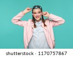 Happy funny girl in casual or hipster style, pigtail hairstyle, standing, holding two blue and pink glasses and looking away with toothy smile, Indoor studio shot, isolated on blue or green background