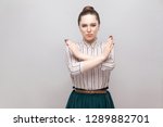 There is no way. Portrait of serious beautiful young woman in striped shirt and green skirt and collected ban hairstyle, standing with block, X sign. indoor studio shot, isolated on grey background.