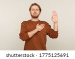 Small photo of I promise to tell truth! Portrait of honest responsible bearded man in sweatshirt standing raising hand and saying swear, making loyalty oath, pledging allegiance. indoor studio shot, gray background