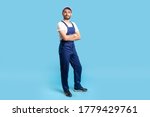 Full length confident professional handyman in overalls standing with crossed hands, smiling at camera. Profession of service industry, house repair. indoor studio shot isolated on blue background