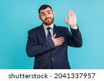 Small photo of I promise to tell truth! Bearded man wearing official style suit standing raising hand and saying swear, making loyalty oath, pledging allegiance. Indoor studio shot isolated on blue background.