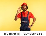 Small photo of Portrait of sappy handyman with beard wearing overalls showing ok sign, being satisfied of done work in time, recommend service industry. Indoor studio shot isolated on yellow background.