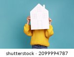 Small photo of Portrait of unknown little kid hiding behind paper house, affordable housing program, advertisement, wearing yellow casual style sweater. Indoor studio shot isolated on blue background.